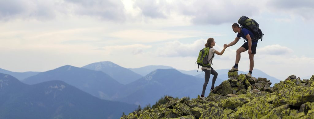 Young tourists with backpacks, athletic man helps slim woman to clime rocky mountain top against